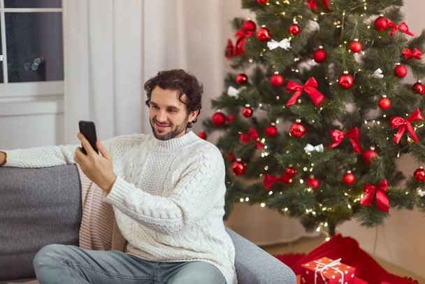 Man Celebration New Year Near Christmas Tree at Home, Video Call or Selfie on Smartphone - 写真・画像
