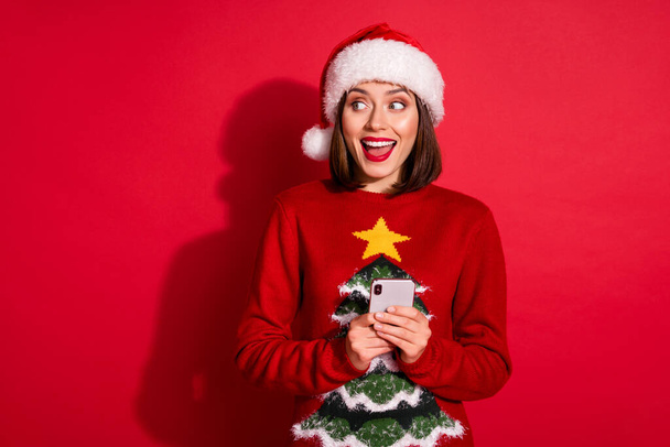 Photo of adorable excited lady dressed pullover celebrating new year chatting gadget looking empty space isolated red color background - Foto, Imagen