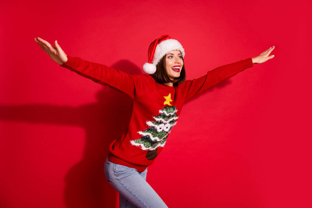 Фото милої дочірньої молодої леді, одягненої в різдвяний принт пуловер, тримає сторони рук, дивлячись на порожній простір ізольований червоний колір фону
 - Фото, зображення