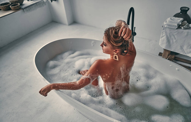 Sexy mujer desnuda posando en el baño blanco sostiene el pelo con las manos arriba disfrutando de la relajación y tomando el sol en la espuma esponjosa. - Foto, imagen