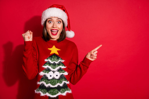 Photo of impressed lucky young woman wear print noel sweater rising fist pointing empty space isolated red color background - Foto, immagini