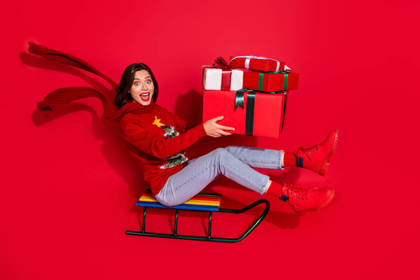 Photo of excited funky young woman wear print sweater riding sleigh delivering new year presents isolated red color background - Foto, Bild