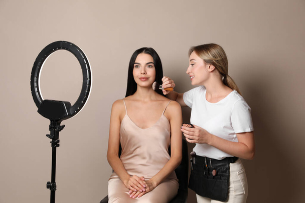 Professional makeup artist working with beautiful young woman against beige background. Using ring lamp - Foto, afbeelding