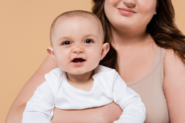 Sonriente morena madre sosteniendo bebé hija aislado en beige  - Foto, Imagen