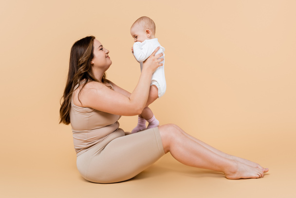 Side view of smiling plus size mother holding child while sitting on beige background  - Photo, Image