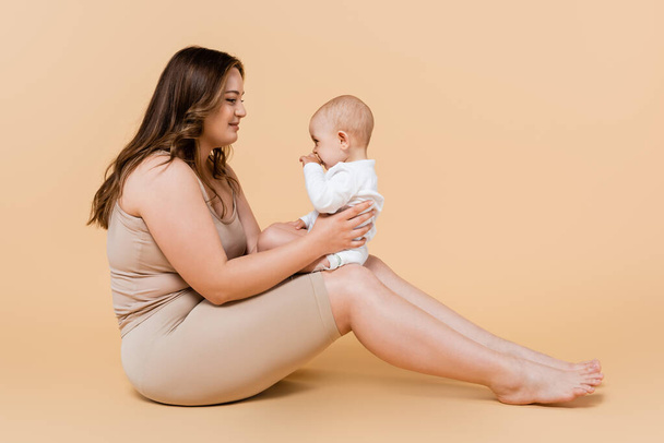 Νέα συν μέγεθος γυναίκα κρατώντας το παιδί, ενώ κάθεται σε μπεζ φόντο  - Φωτογραφία, εικόνα