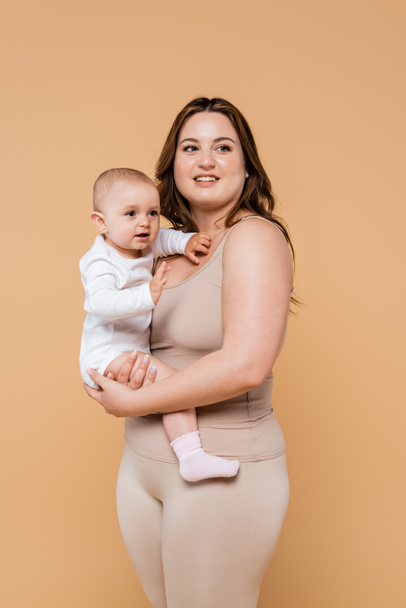 Szczęśliwa kobieta z nadwagą trzymając dziecko i patrząc odizolowany na beżu  - Zdjęcie, obraz