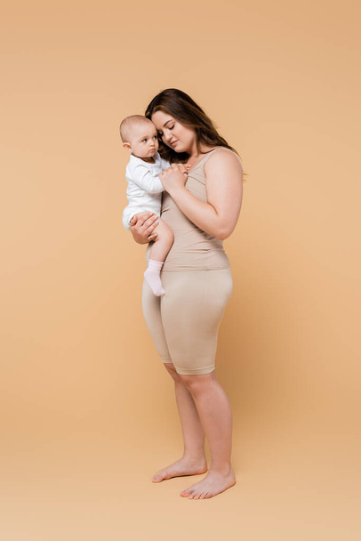 Full length of plus size mother holding baby daughter on beige background  - Foto, Imagem