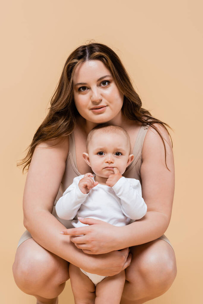 Young plus size woman looking at camera while hugging child isolated on beige  - Fotó, kép
