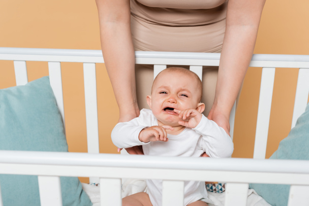 Plus size mother standing near crying baby daughter in crib isolated on beige  - Valokuva, kuva