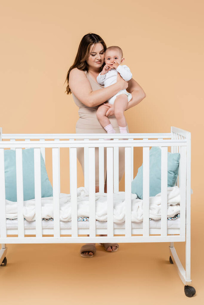 Young plus size woman holding baby near crib on beige background  - Zdjęcie, obraz