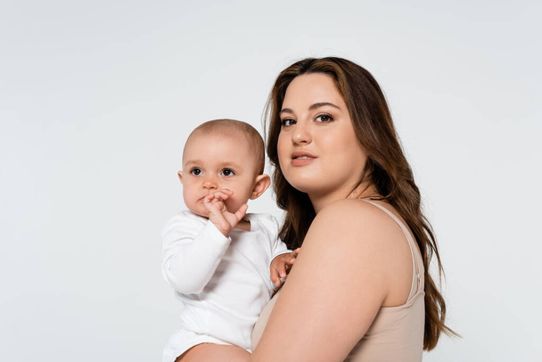 Pretty body positive woman hugging baby girl and looking at camera isolated on grey - Foto, afbeelding