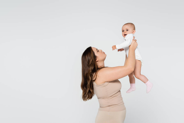 Giovane madre con labbra da broncio in sovrappeso mentre solleva bambino isolato su grigio - Foto, immagini