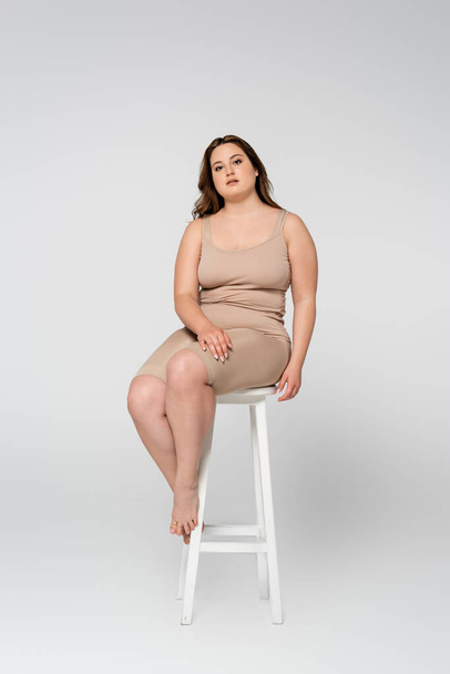 Pretty plus size woman looking at camera while sitting on chair on grey background - Foto, afbeelding