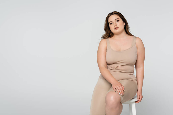 Brunette body positive woman sitting on chair and looking at camera isolated on grey - Fotografie, Obrázek