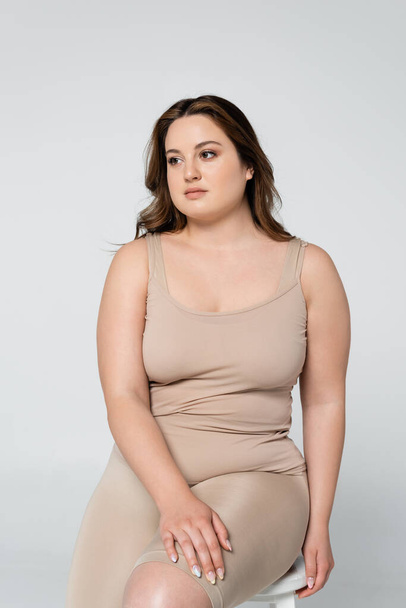 Pretty body positive woman looking away while sitting on chair isolated on grey - Foto, imagen
