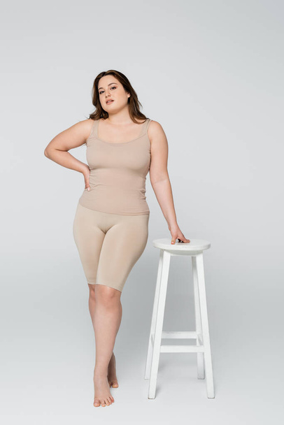 Full length of pretty plus size woman holding hand on hip near chair on grey background - Photo, image