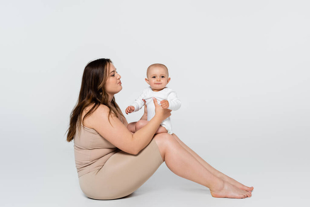 Mladá žena s nadváhou drží dítě, zatímco sedí na šedém pozadí - Fotografie, Obrázek
