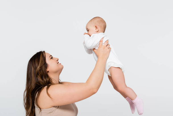Side view of happy plus size mother lifting baby isolated on grey - Photo, Image