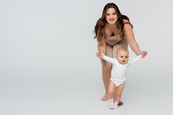 Happy plus size woman looking at camera while holding hands of baby girl on grey background - Fotó, kép