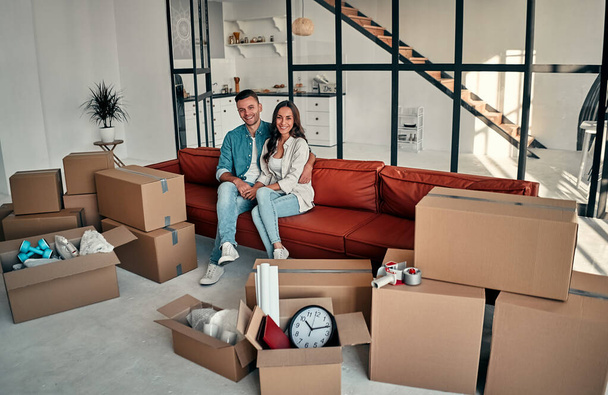 Young married couple sitting on couch in living room at house. Smiling happy wife and husband relaxing resting unopened belongings still in their cardboard boxes. Moving and relocate new home concept. - Photo, Image