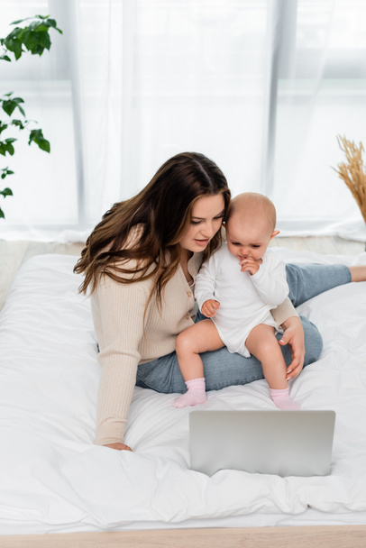 Mulher com sobrepeso segurando bebê filha perto de laptop na cama  - Foto, Imagem