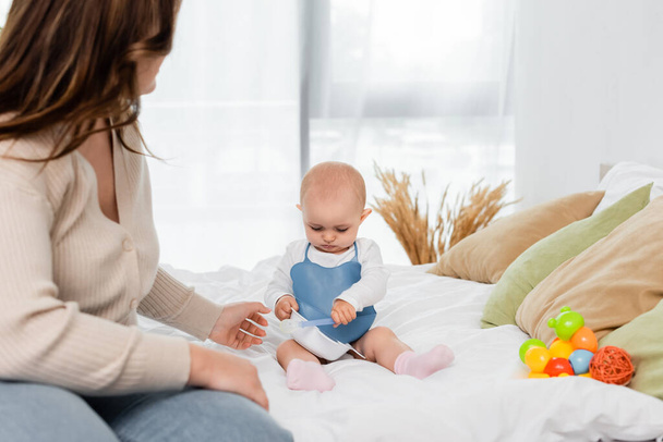 Giovane madre positiva del corpo seduta vicino al bambino con piatto e cucchiaio sul letto  - Foto, immagini