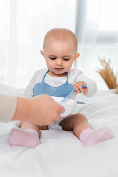 Woman holding plate with baby food near daughter with spoon on bed  - Foto, Imagen