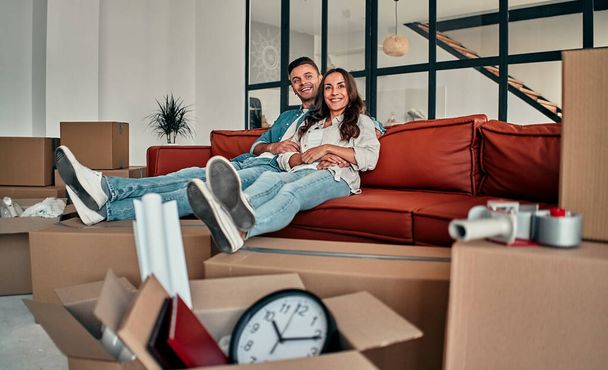 Fiatal házaspár ül otthon a kanapén a nappaliban. Boldog férj és feleség jól érzik magukat, alig várják, hogy új otthonuk legyen. Költözés, házvásárlás, lakás koncepció. - Fotó, kép
