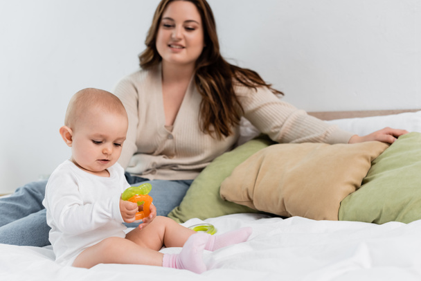Baby girl playing with toys near blurred plus size mother on bed  - Foto, imagen