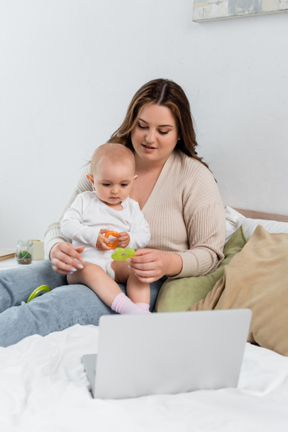 Νεαρή θετική στο σώμα γυναίκα κρατώντας παιχνίδι κοντά στο μωρό και φορητό υπολογιστή στο κρεβάτι  - Φωτογραφία, εικόνα