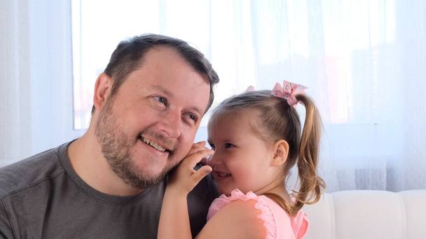 little daughter, 3 years old, tells her dad a secret, play together at home. Fathers day. Handsome young man at home with his little cute girl are having fun together. Fatherhood concept, happy - Photo, Image