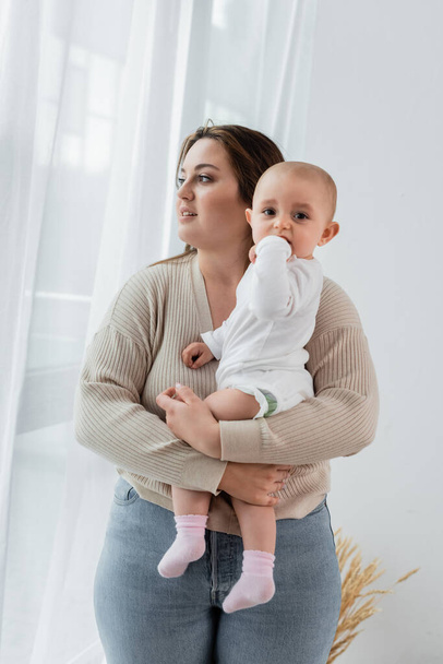 Morena cuerpo positivo mujer sosteniendo bebé niña cerca de cortinas en casa  - Foto, imagen