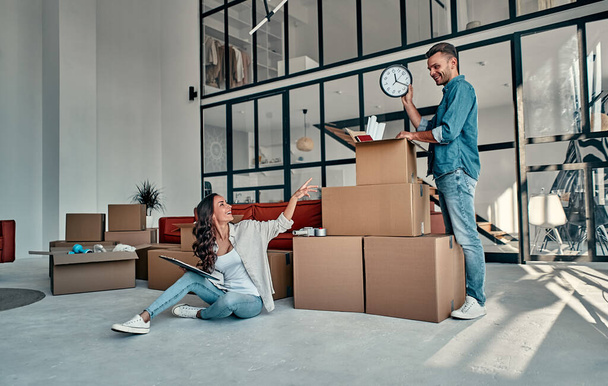 A young married couple in the living room in the house unpack boxes with things. A woman with a folder checks if everything is in place. Moving, buying a house, apartment concept. - Fotó, kép
