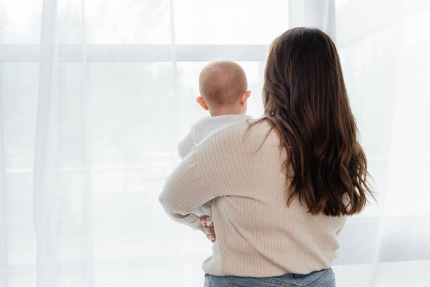 Back view of plus size woman holding baby near window at home  - Photo, Image