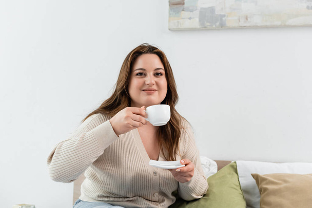 Pleased body positive woman holding cup and saucer on bed at home  - Фото, зображення