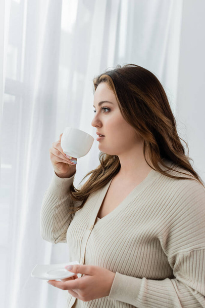 Молодая женщина plus size, держащая чашку кофе у штор дома  - Фото, изображение
