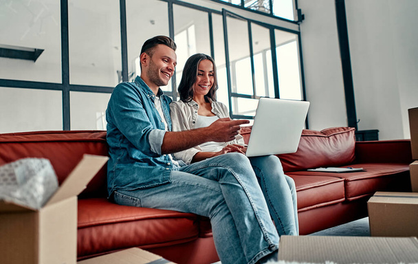 新しい家に移動しながら、コンピュータ無線インターネットを使用して一緒に床に座っている若いカップル。引っ越し、家を買う、アパートのコンセプト. - 写真・画像
