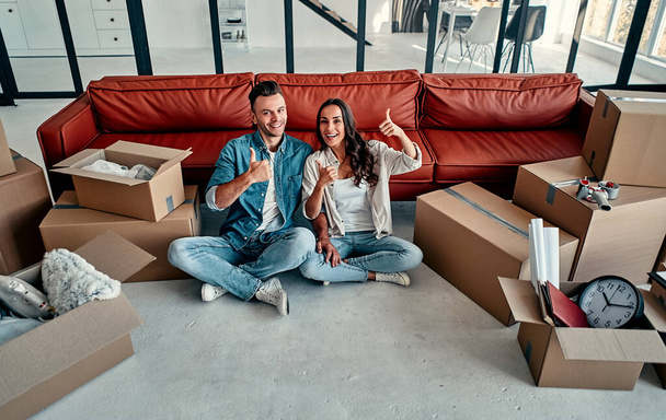 Young married couple show thumb up in living room at house. Smiling happy wife and husband relaxing resting unopened belongings still in their cardboard boxes. Moving and relocate new home concept. - Photo, Image