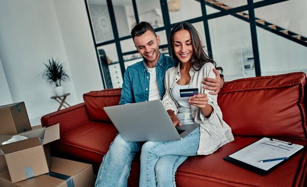 Nuori pari istuu lattialla yhdessä tietokoneen langattoman internetin ja luottokortin avulla muuttaessaan uuteen kotiin. Muutto, talon ostaminen, asunnon käsite. - Valokuva, kuva