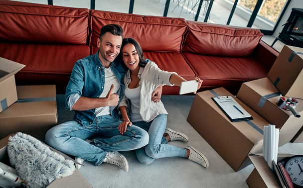 Young couple taking selfies with her smartphone in their new home. Moving, buying a house, apartment concept. - Photo, Image