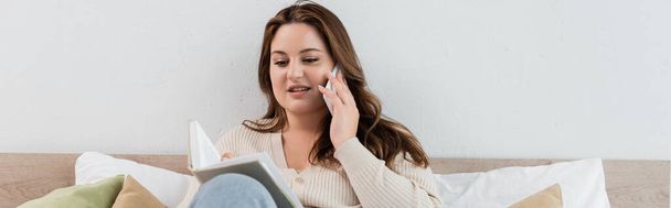 Positive plus size woman talking on cellphone and reading book in bedroom, banner  - Фото, зображення