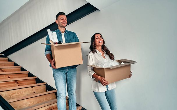 Moving to a new home. Happy young couple holding cardboard boxes while going down the stairs. - Photo, Image