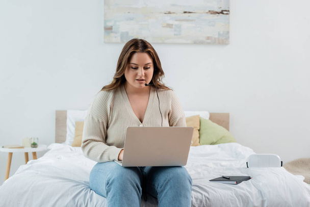 Freelancer with overweight using laptop and headset near notebook on bed  - Photo, Image
