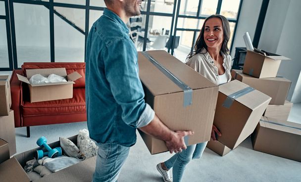 A young married couple in the living room in the house unpack boxes with things. Happy husband and wife are having fun, are looking forward to a new home. Moving, buying a house, apartment concept. - Photo, Image