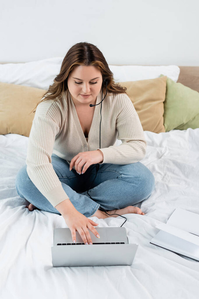 Body positive teleworker in headset using laptop near notebook on bed  - Foto, imagen