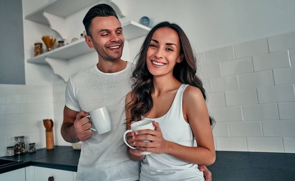 Szczęśliwa młoda para trzyma filiżanki kawy stojąc razem w kuchni. - Zdjęcie, obraz