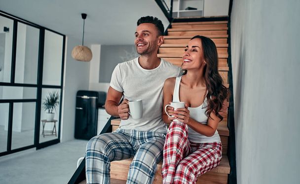 パジャマを着た若いカップルが家の階段に座って朝のコーヒーを飲んでいます. - 写真・画像
