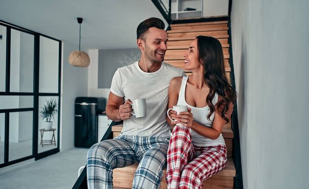 Una joven pareja enamorada en pijama está sentada en las escaleras de su casa y tomando café de la mañana.. - Foto, imagen