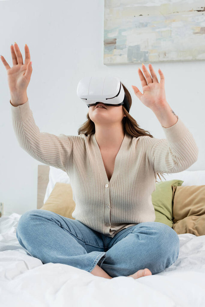 Young body positive woman gaming in vr headset while sitting on bed  - Foto, Imagen